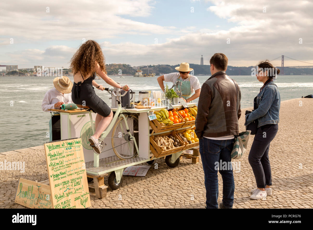 Young women rides bike to make smoothie, while tourist watch in Lisbon Portugal along the Tagus River.  Fresh smoothie stand Stock Photo