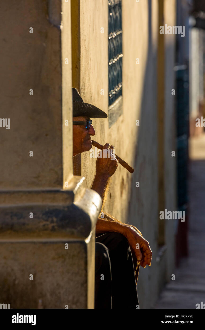 An old Cuban smokes a cigar and sits on a staircase in the warm light of the sunset, Trinidad, Cuba, Sancti Spíritus, Cuba Stock Photo