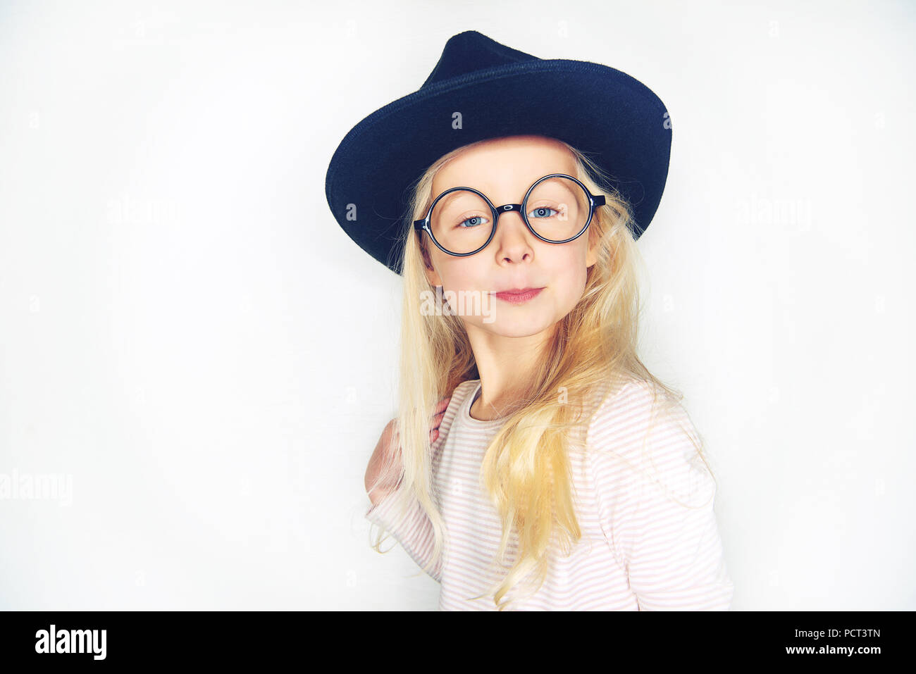 Cute little girl with long blonde hair wearing a black hat and glasses smiling while standing against a white background Stock Photo