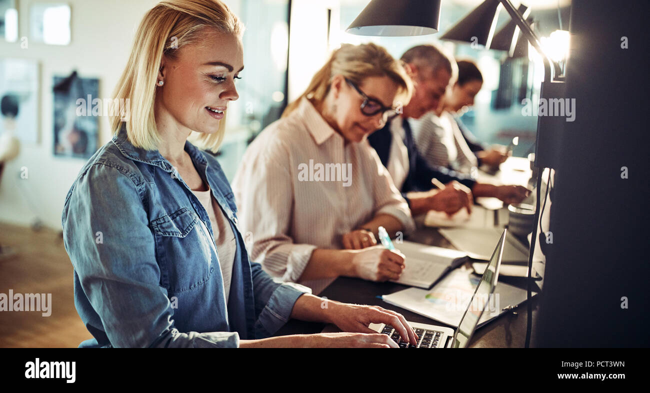 Smiling young female designer working on a laptop while sitting in a row with colleagues at a table in an office Stock Photo
