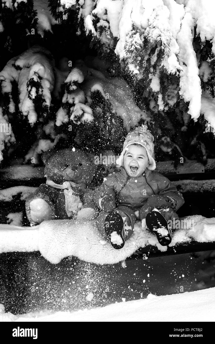 Little boy sitting with teddy bear on a bench in the snow and laughing Stock Photo