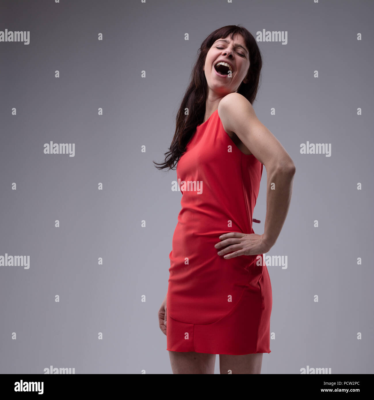 Happy woman in red dress standing singing with her hand to her hip and a diva style gesture over grey with copy space Stock Photo