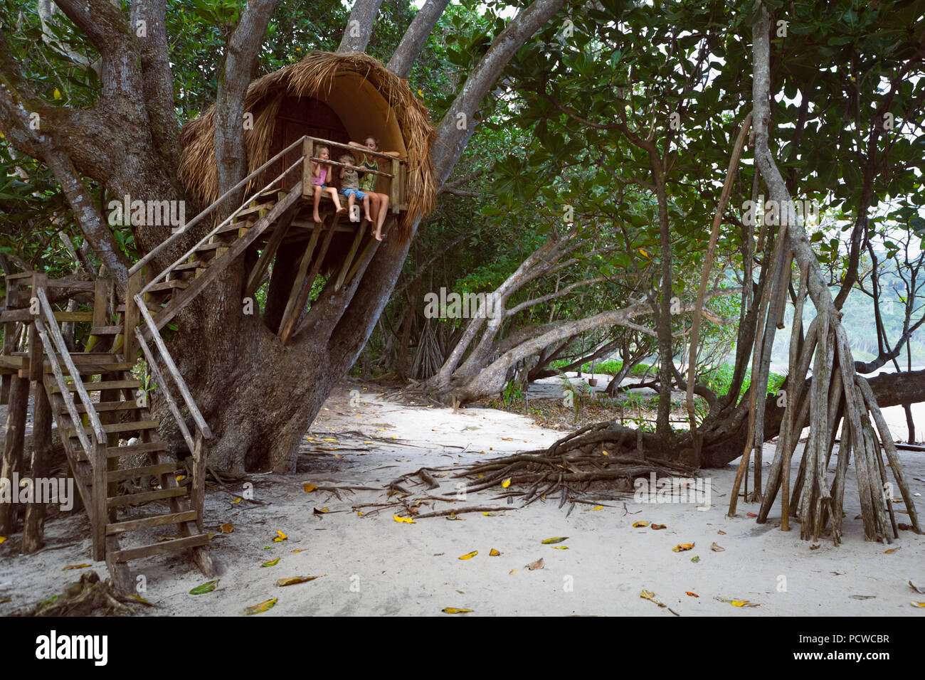 Happy children have fun on family holidays walk. Kids, mother sit on balcony edge of tree house dangling legs, look at sea beach through jungle. Stock Photo