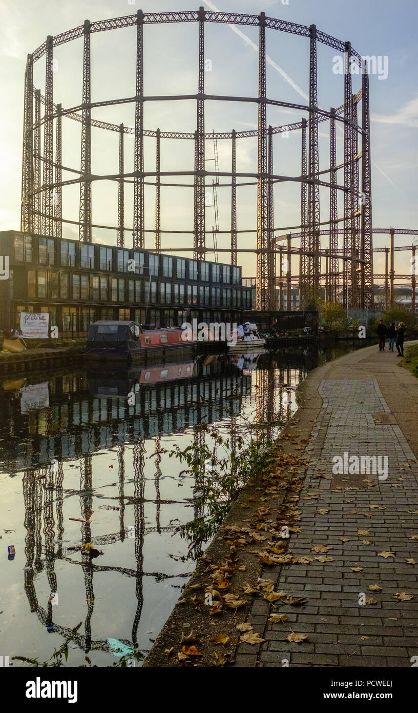 Old Industrial gas holders with modern buildings and Barge, reflecting in Regents Canal Hackney during autumn with leaves on walkway, London, England. Stock Photo