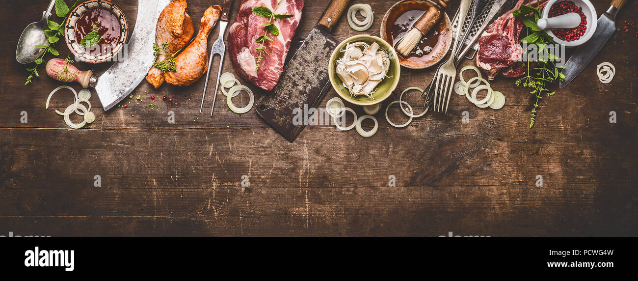 Various grill an bbq meats on rustic wooden background with aged kitchen and butcher tools, herbs, spices, seasoning and sauce, top view, border Stock Photo