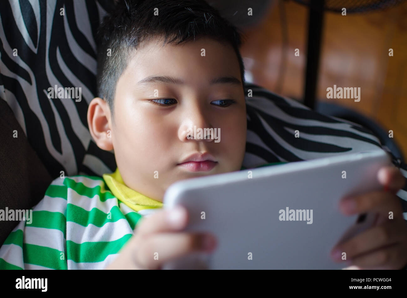 A young boy, new generation kid lying down in a sofa playing a game on a tablet computer at home. Stock Photo