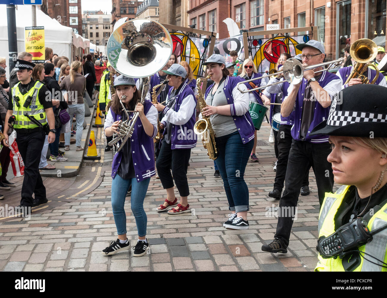 Glasgow, Scotland, UK. 04th August 2018. Police officers accompany participants in the Carnival Procession of the Merchant City Festival. Shown are the Baybeat Street band, and behind them people carrying 'stained glass windows' of the Mackintosh Rose. The festival is part of Festival 2018 a city-wide cultural event running in parallel with Glasgow 2018, the European Championships. Credit: Elizabeth Leyden/Alamy Live News Stock Photo