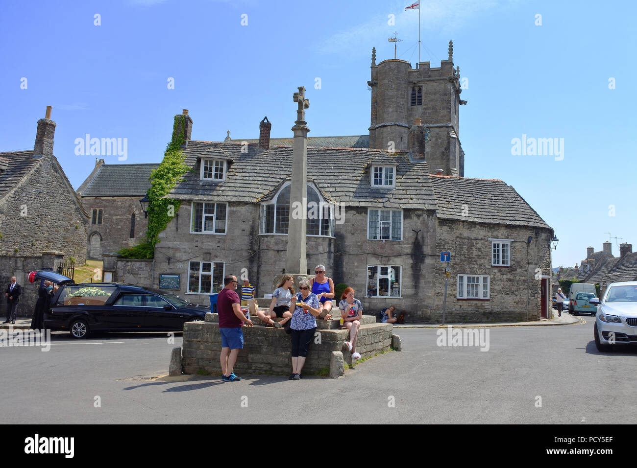 Tourist family eating food in the square at Corfe with a funeral in progress in the background - John Gollop Stock Photo