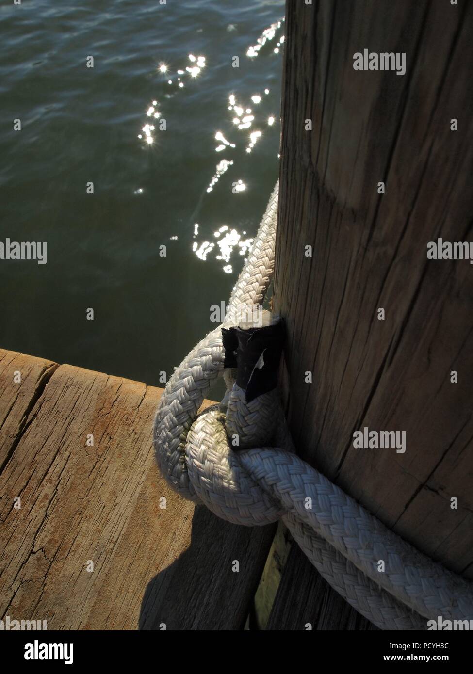 Close-up of thick braided rope tied in knot around wooden dock piling with light reflections glistening in water. Unique perspective of nautical scene Stock Photo