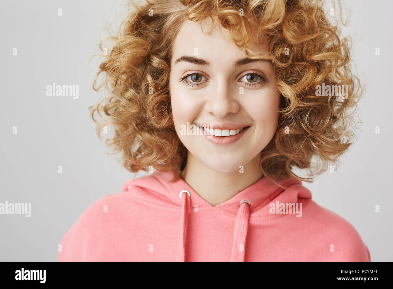 Ready To Help You And Show Around Pleasant Good Looking Girl With Curly Hair Smiling Happily While Standing Over Gray Background In Stylish Hoodie C Stock Photo Alamy