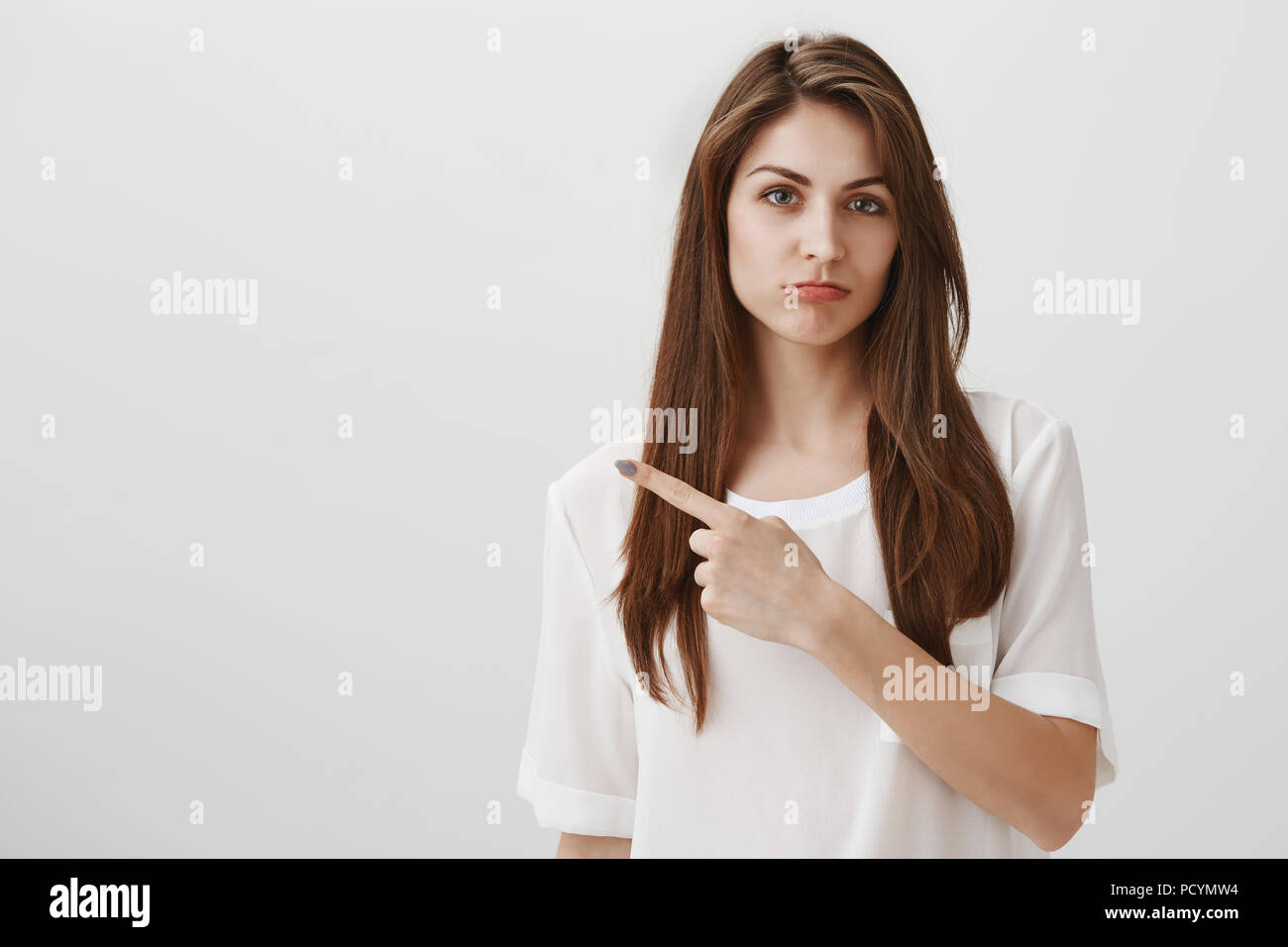 Extremely jealous girlfriend indicating at purse she wants as present. Portrait of attractive brunette girl pointing left and sulking, expressing regr Stock Photo