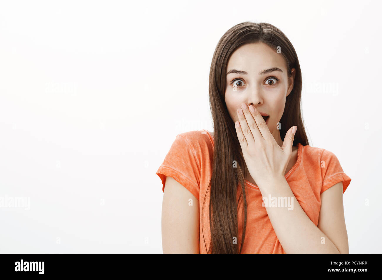 Chatty girl likes gathering rumors in office. Portrait of surprised and shocked attractive female model in orange t-shirt, covering opened mouth with  Stock Photo