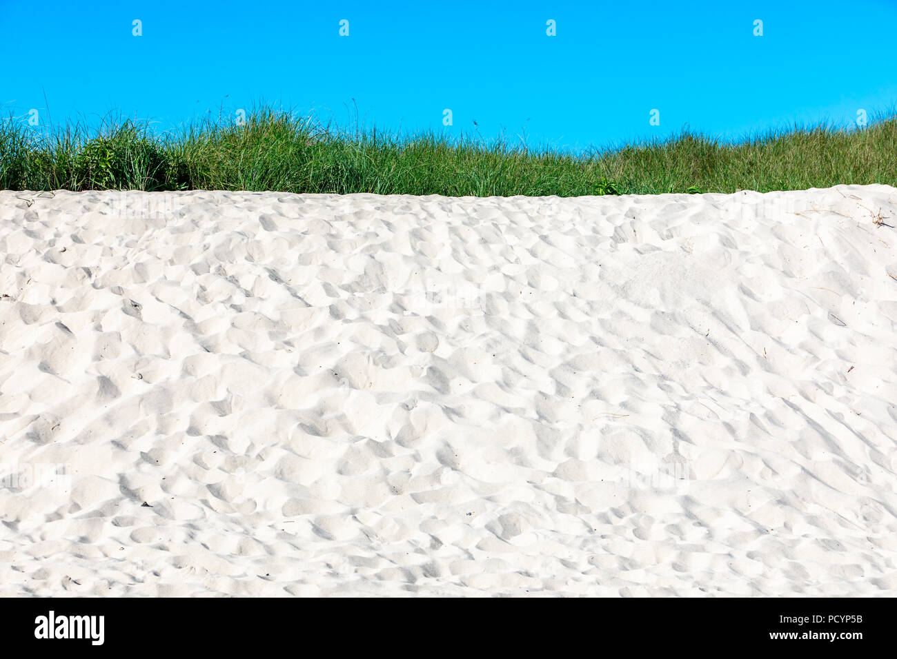 detail shot of a sandy dune with a small piece of grass at the top and a small section of blue sky Stock Photo