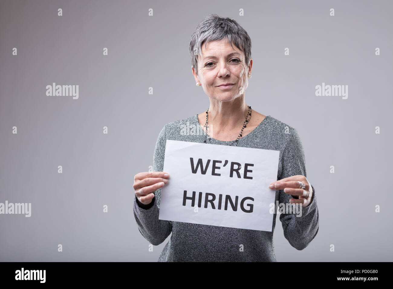 Personnel manageress holding up a sign - We're Hiring - in front of her chest in an employment and human resources concept over grey with copy space Stock Photo