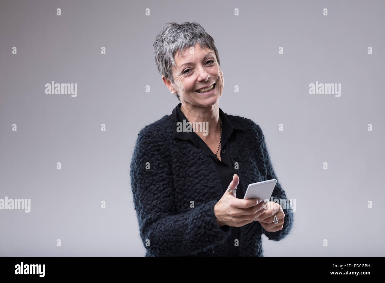 Charismatic older woman with a sweet smile holding a mobile phone in her hands as she looks at the camera over grey with copy space Stock Photo