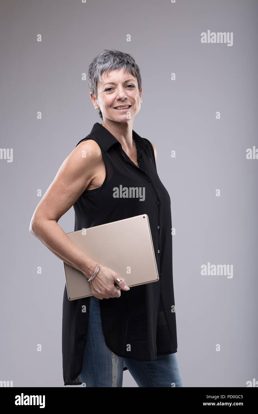 Trendy attractive older woman holding a tablet-pc under her arm as she stands smiling at the camera in a side view isolated on grey Stock Photo