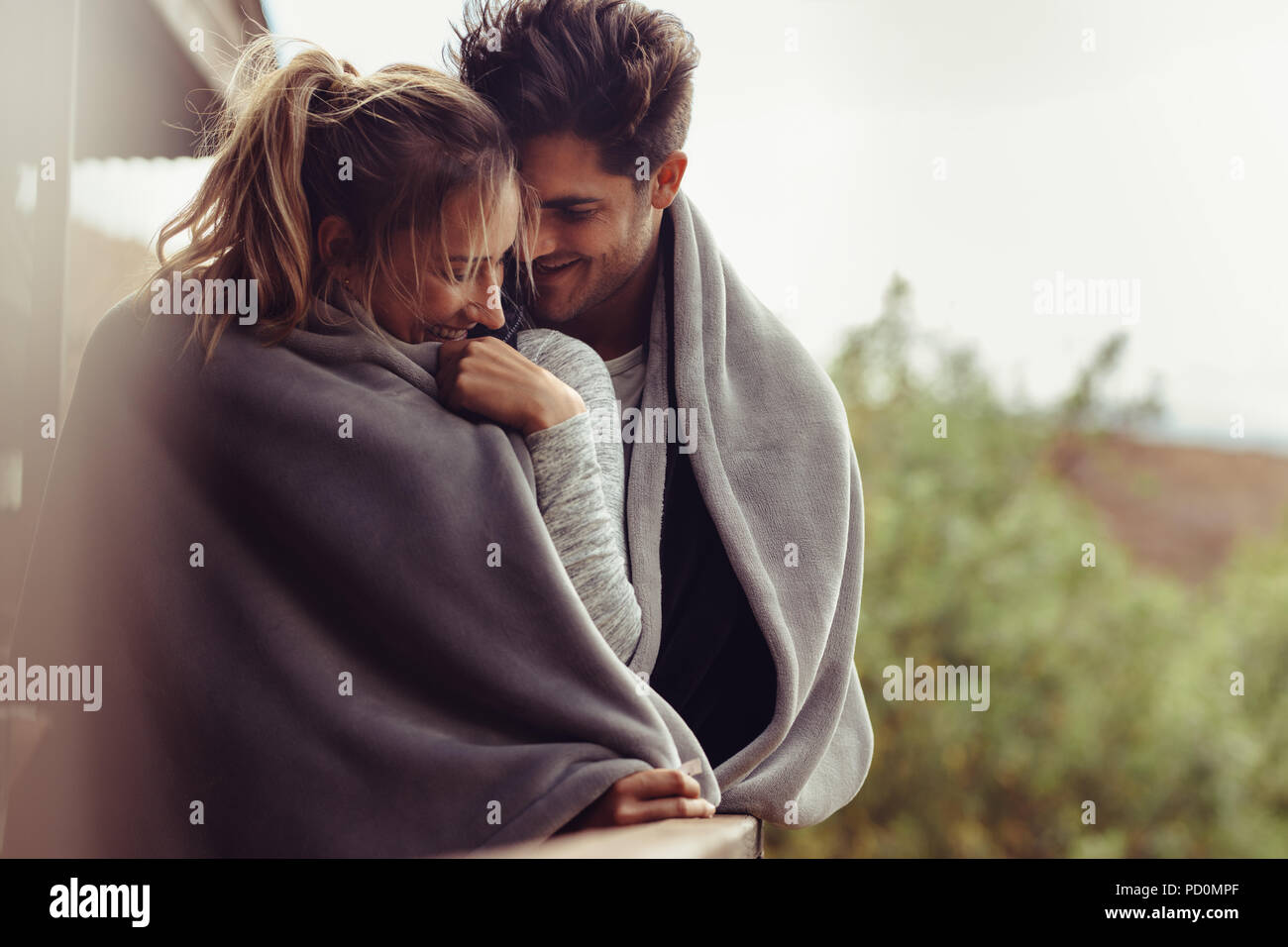 Romantic couple on a winter holiday. Man and woman standing together in a hotel room balcony wrapped in blanket. Couple embracing and smiling. Stock Photo