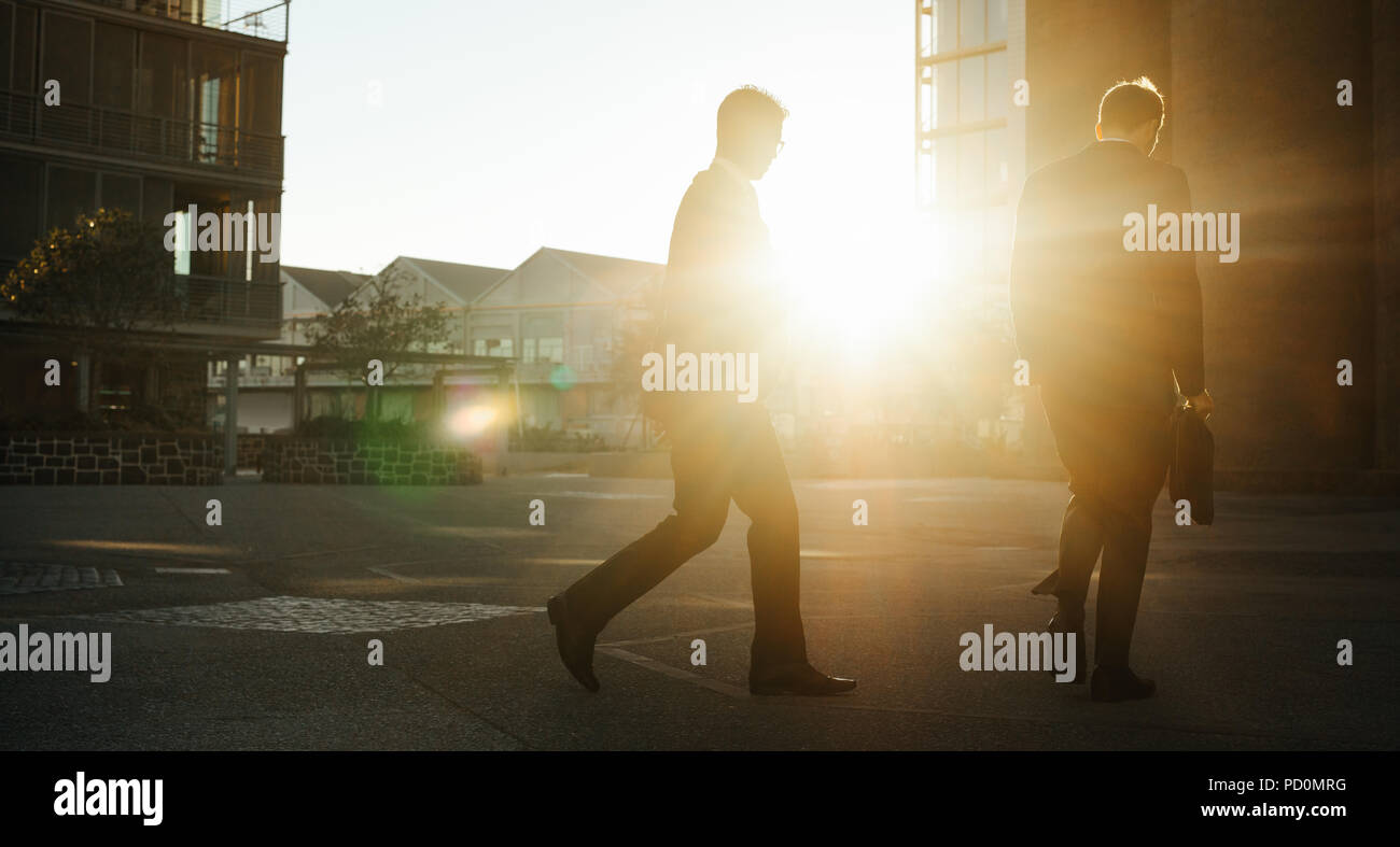 Men commuting to office early in the morning with rising sun in the background. Businessmen with office bag in hand walking on the street to office. Stock Photo