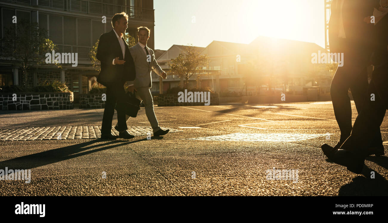 Men in formal clothes commuting to office early in the morning carrying office bags. Business colleagues talking while walking on city street with sun Stock Photo
