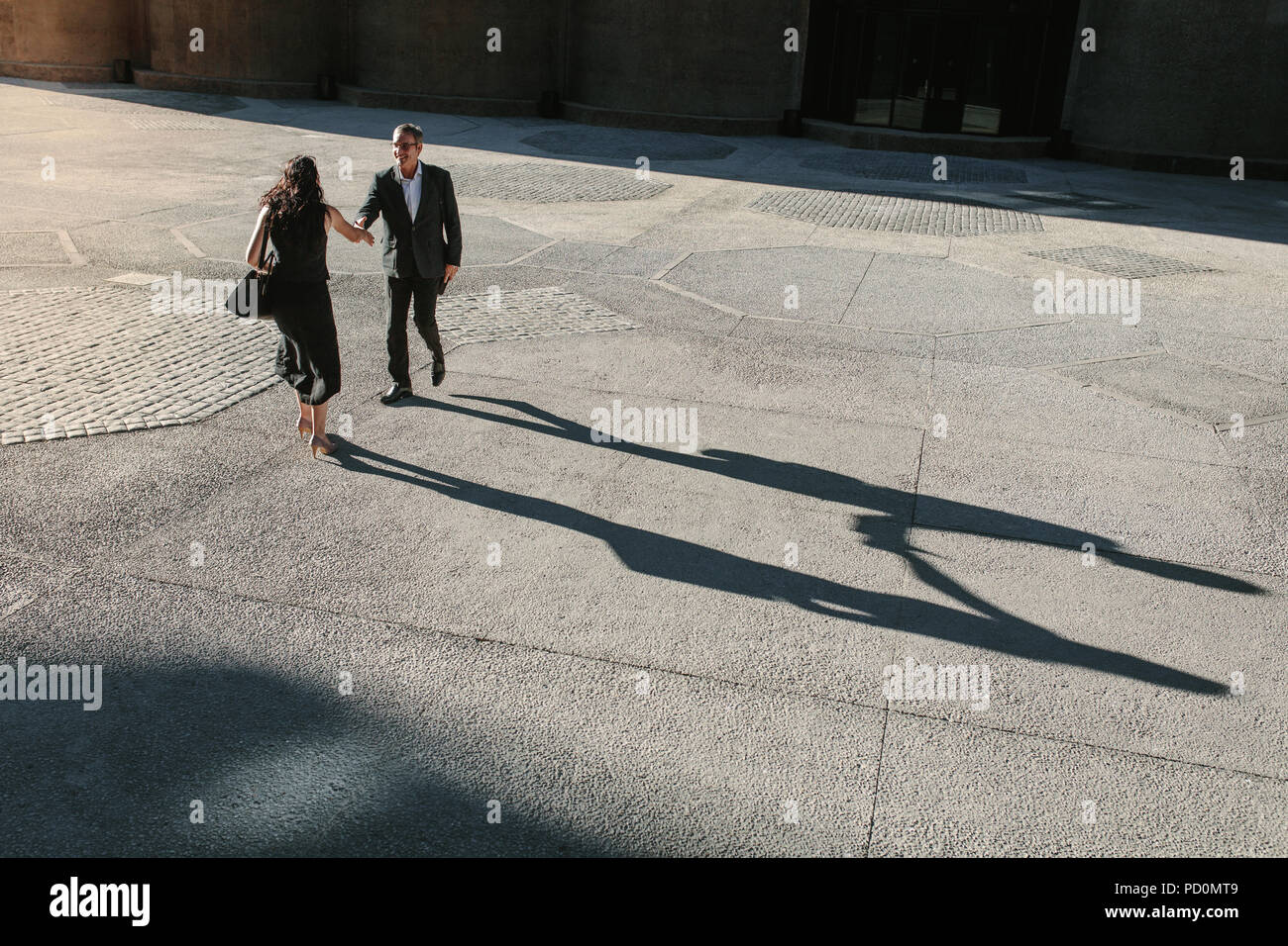 Businesswoman and a businessman walking forward to shake hands on a street with their long shadows on the ground. Business people greeting each other  Stock Photo