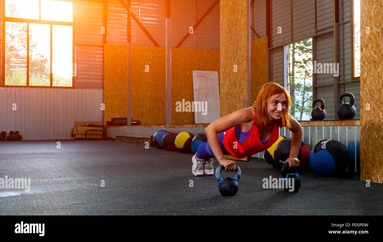 A young red-haired woman in a pink top and purple leggings doing push-ups, hands on weights in a sports gym. Push-up technique with weight Stock Photo