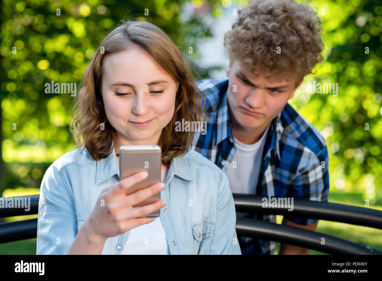 Beautiful brunette girl in summer in a park on nature. In hands of holding a smartphone communicates in social networks. A young man spies on her sms. The concept of distrust problem in relationship. Stock Photo
