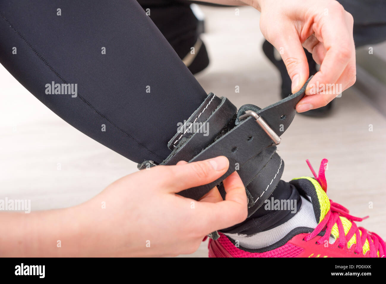 Young woman in trainers buckling a leather strap to her ankle as she sits on the floor of a gym
