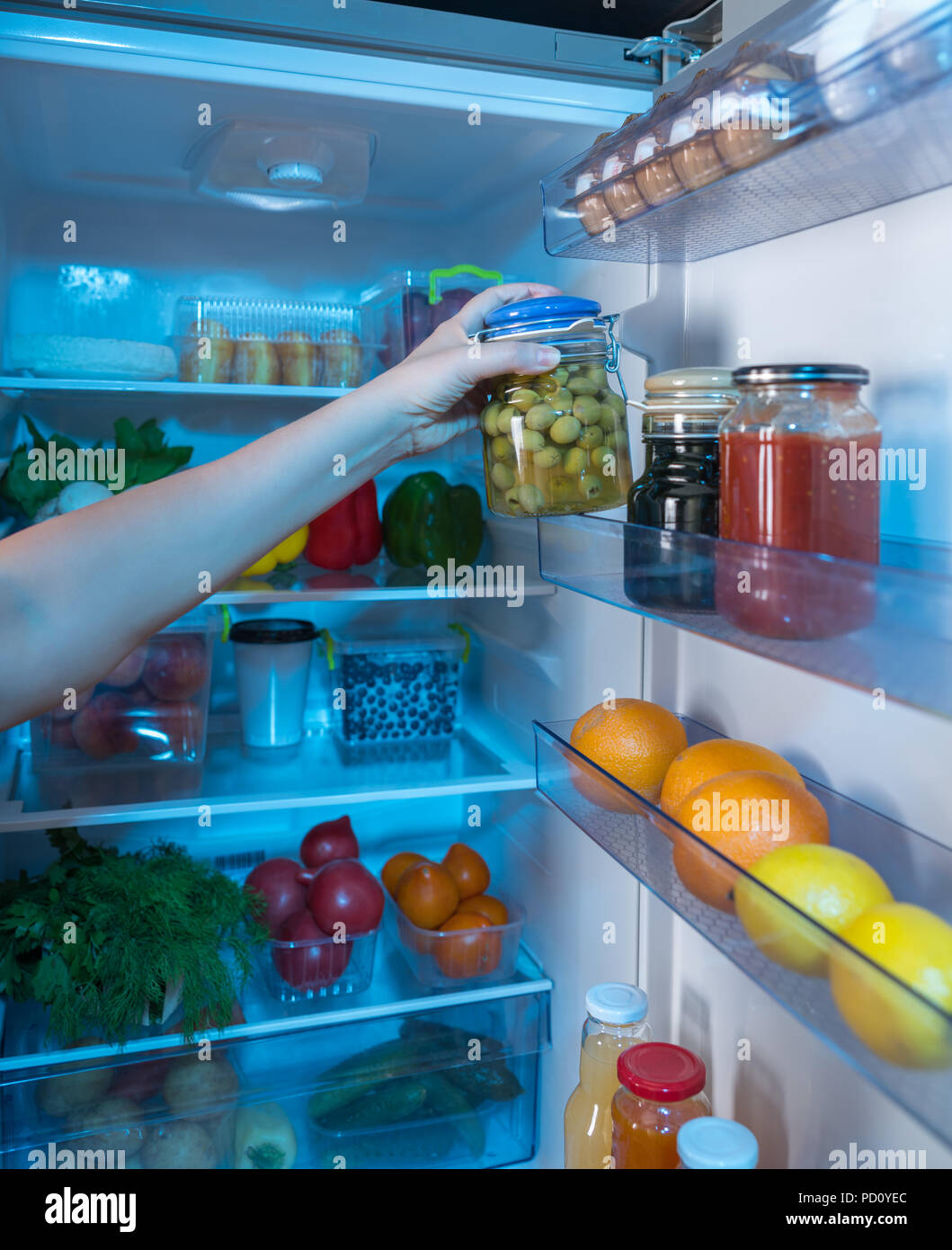 Person reaching for jar with green olives in open fridge full of food