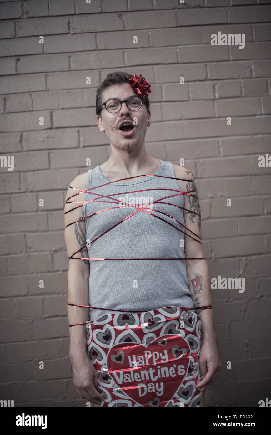 Young white man with tattoos wrapped in a red gift ribbon, with a red bow in the side of the head, curly moustache, and vintage glasses. Stock Photo