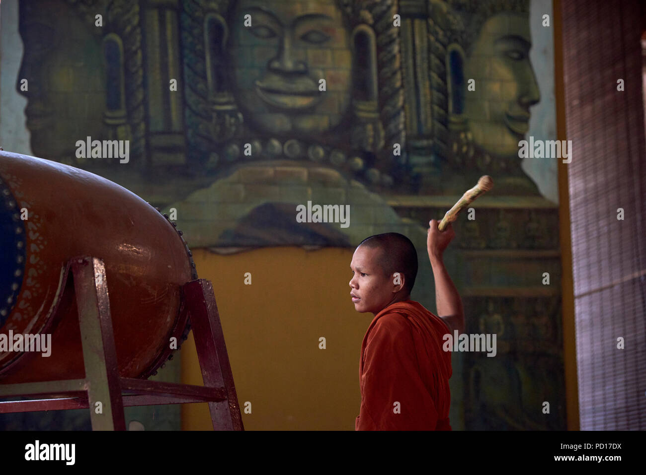 A young Buddhist monk playing a tambour during a ceremony at a monastery in Siem Reap, Cambodia, with faces of gods painted in a mural in the backgrou Stock Photo