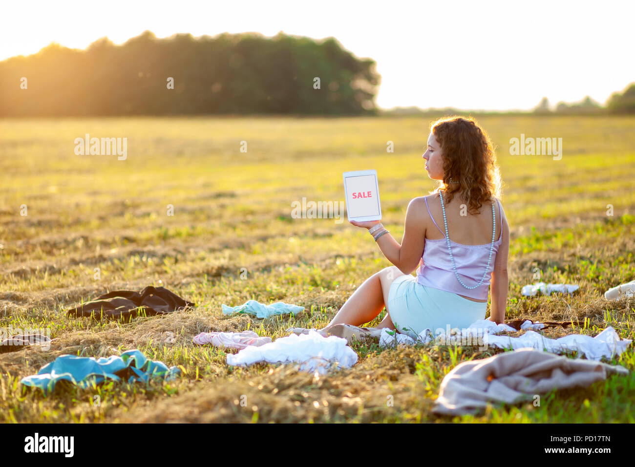 Dressy woman sitting in the middle of the field between scattered cloth and holding a tablet with SALE title during sunset. Fast fashion over-producti Stock Photo
