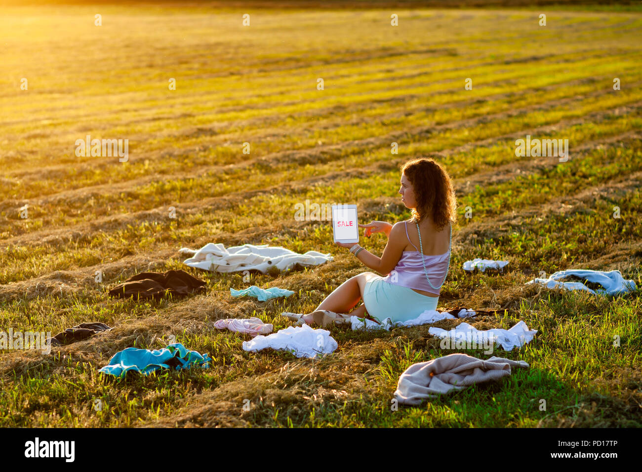 Dressy woman sitting in the middle of the field between scattered cloth and holding a tablet with SALE title during sunset. Fast fashion over-producti Stock Photo