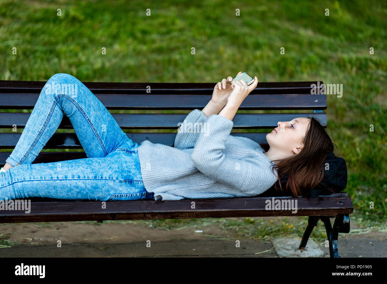 Girl schoolgirl. In summer, he lies on a bench in city. In the hands of  holding a smartphone, watching a video. Concept of rest after school.  Emotion is a pleasure pleasure Stock