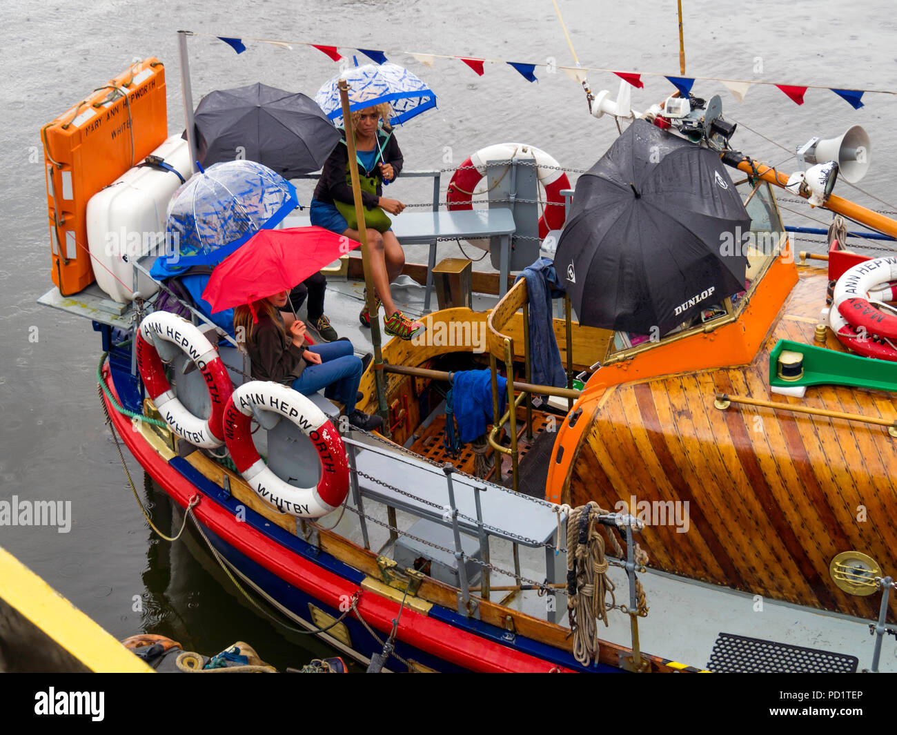 Passengers on the old Whitby Lifeboat 'Mary Ann Hepworth' in Whitby harbour waiting to go on a summer pleasure trip  umbrellas up in heavy rain Stock Photo