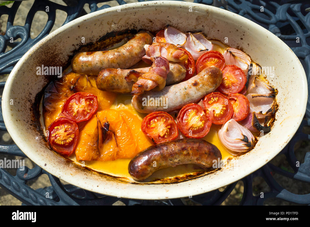Oven roasted Pork Sausages bacon yellow peppers tomatoes and onions in an oval dish on an outside table ready for serving alfresco Stock Photo