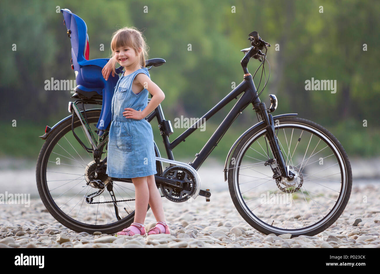 Small pretty blond girl in blue dress standing on pebbles in front of modern bicycle with child seat on blurred green trees background on summer day.  Stock Photo