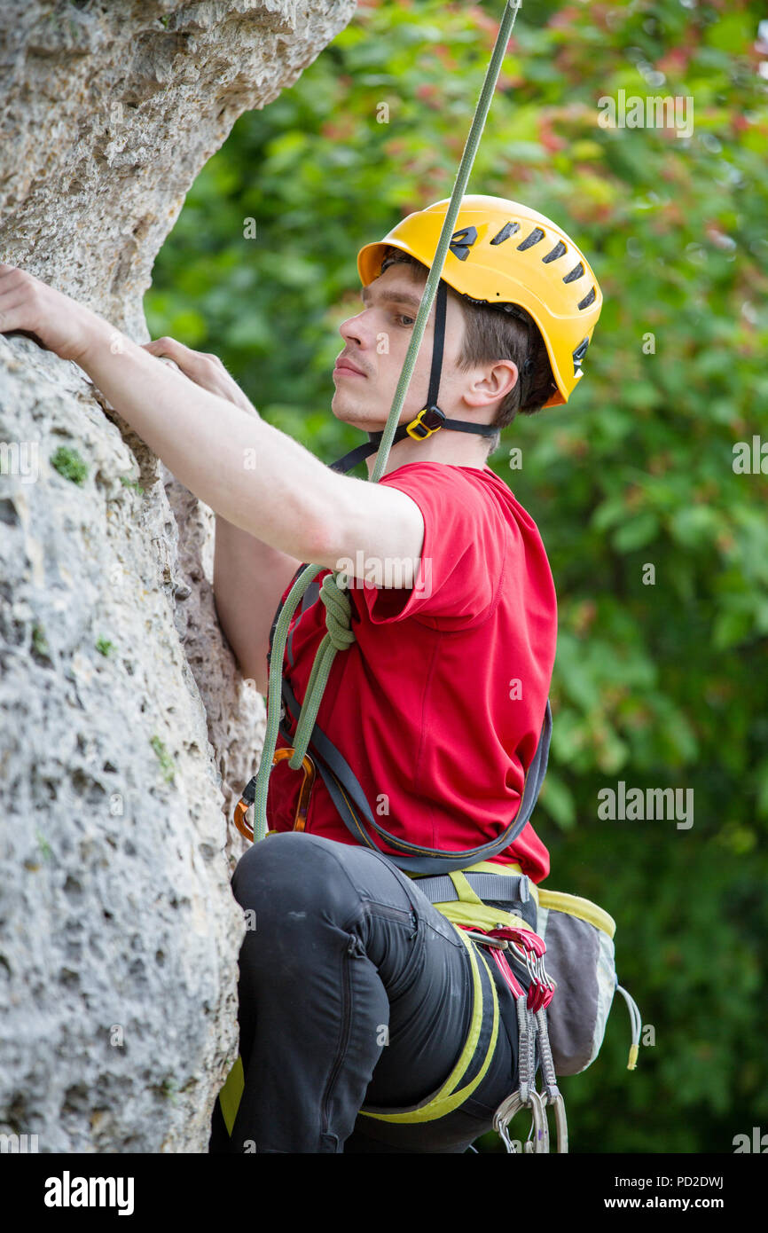Photo of sportsman in yellow helmet clambering over rock against background of green trees Stock Photo