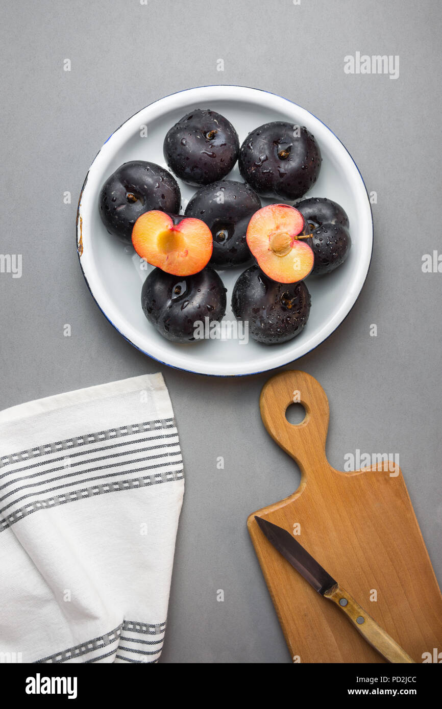 Dark red big plums whole and halved on white vintage enamel plate. Wooden cutting board knife white cotton towel on gray stone background. Minimalist  Stock Photo