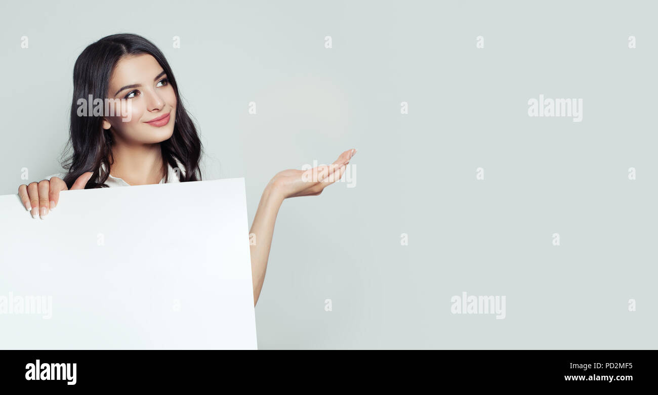 Smiling business woman showing empty open hand and holding white blank paper banner background. Young woman portrait, business and education concept Stock Photo