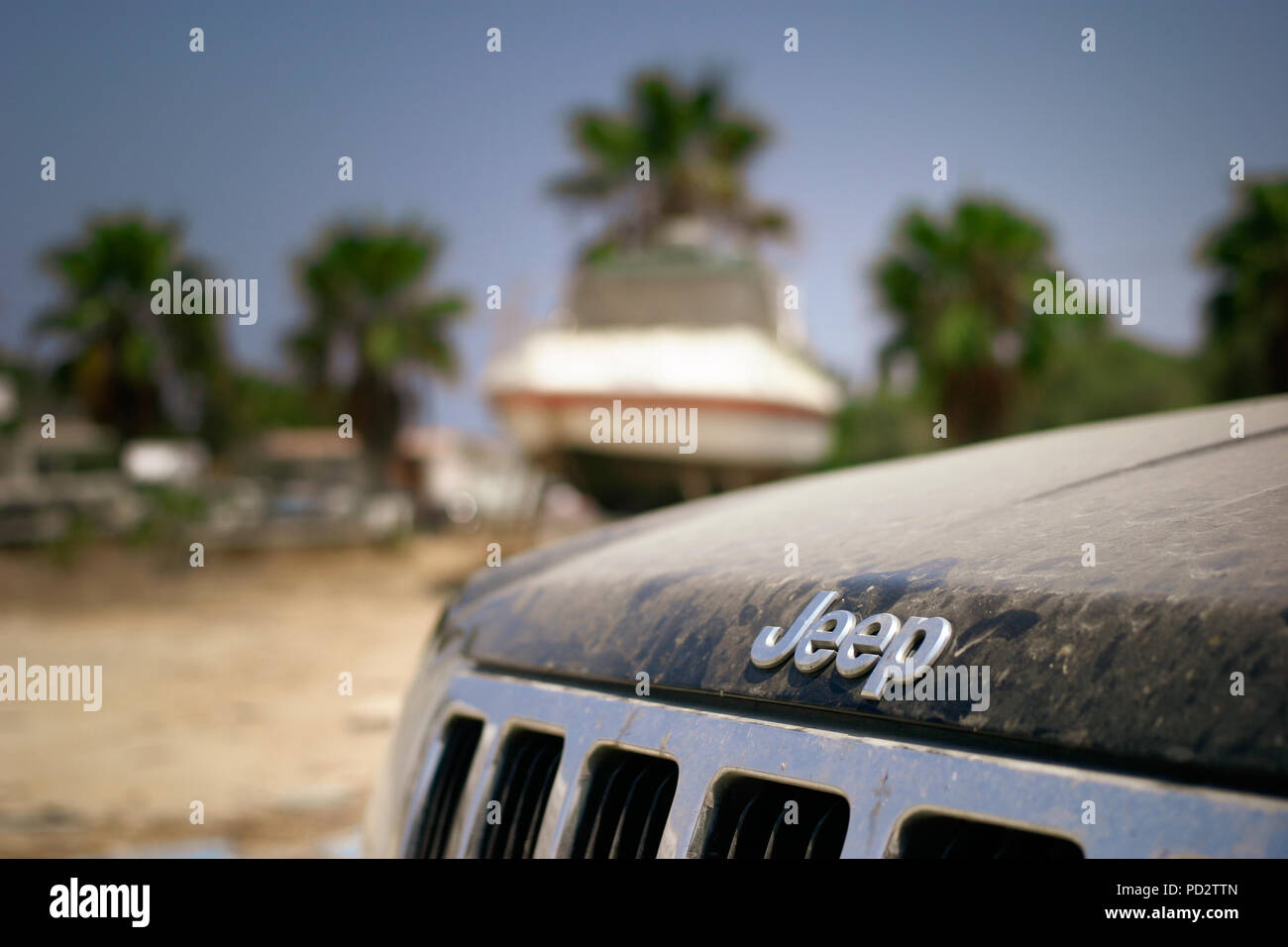 A dusty jeep parked in front of a boat storage yard, close up of jeep branding and the dirt on the bonnet. Boat in the background, with palm trees. Stock Photo