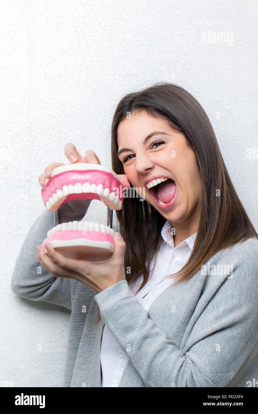 Close up fun portrait of attractive young girl holding oversize human teeth prosthesis.Woman with open mouth against light textured background. Stock Photo