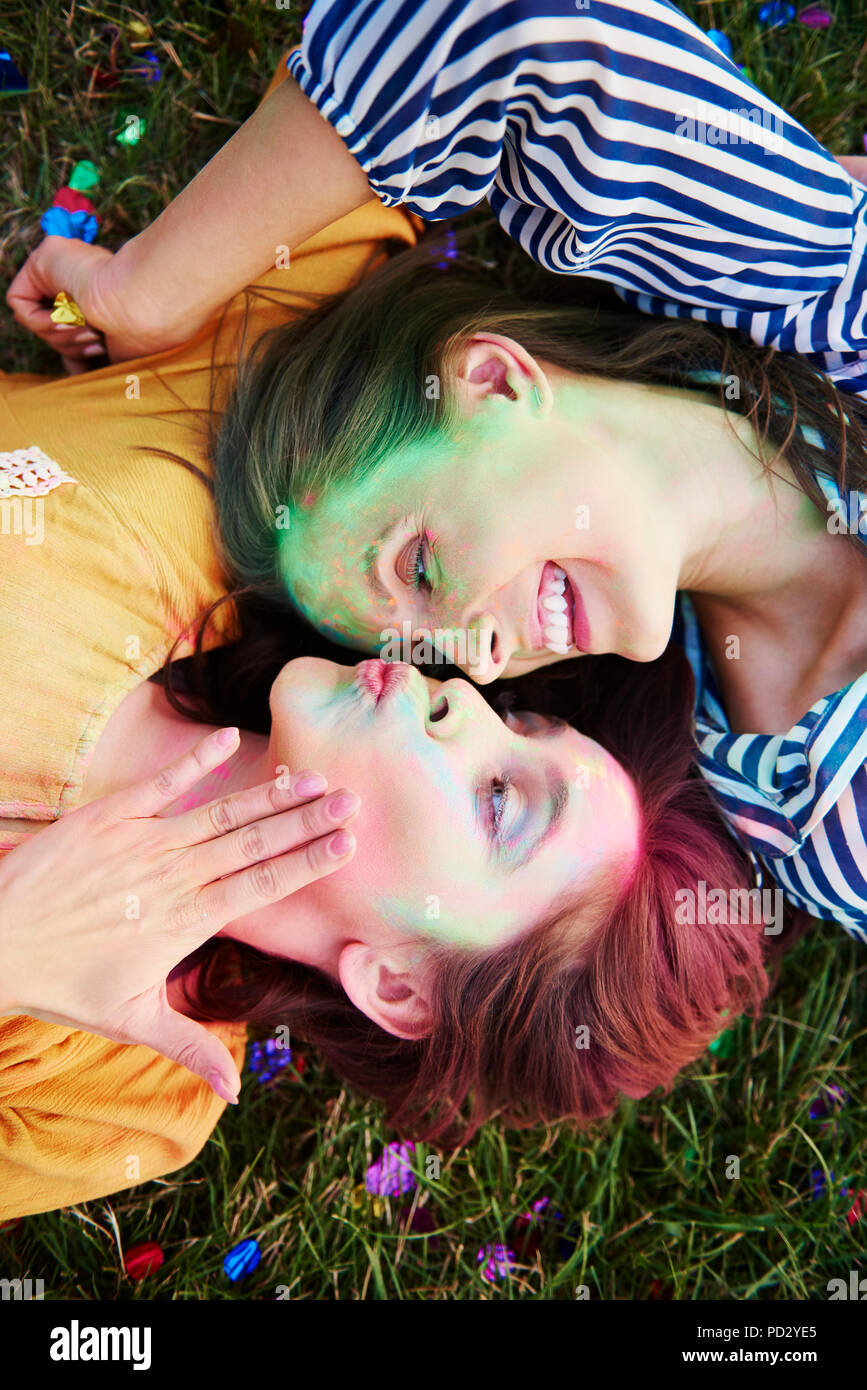 Two young women covered in coloured chalk powder lying on grass at Holi Festival, overhead view Stock Photo