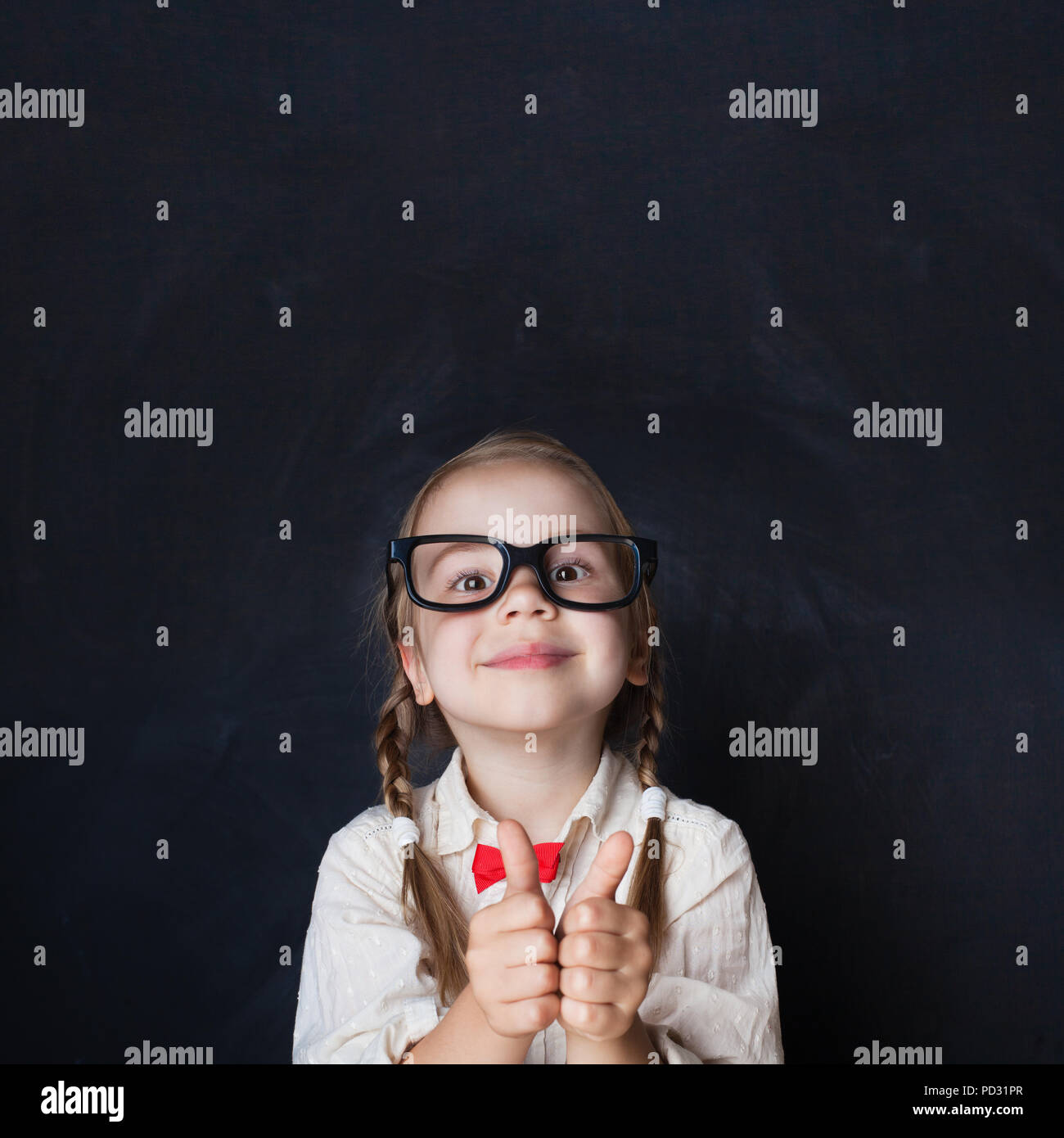 Сurious Girl in Glasses Showing Hands with Thumb Up and Smiling on Chalk Board Background. Child Thumb Up. Back to school, Creativity and Education Co Stock Photo