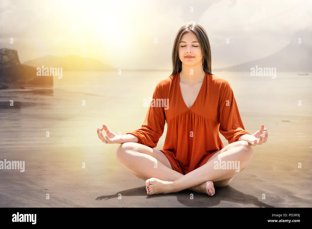 Portrait of young woman meditating at riverside. Girl sitting with eyes closed on sand against dreamy sunset background. Stock Photo