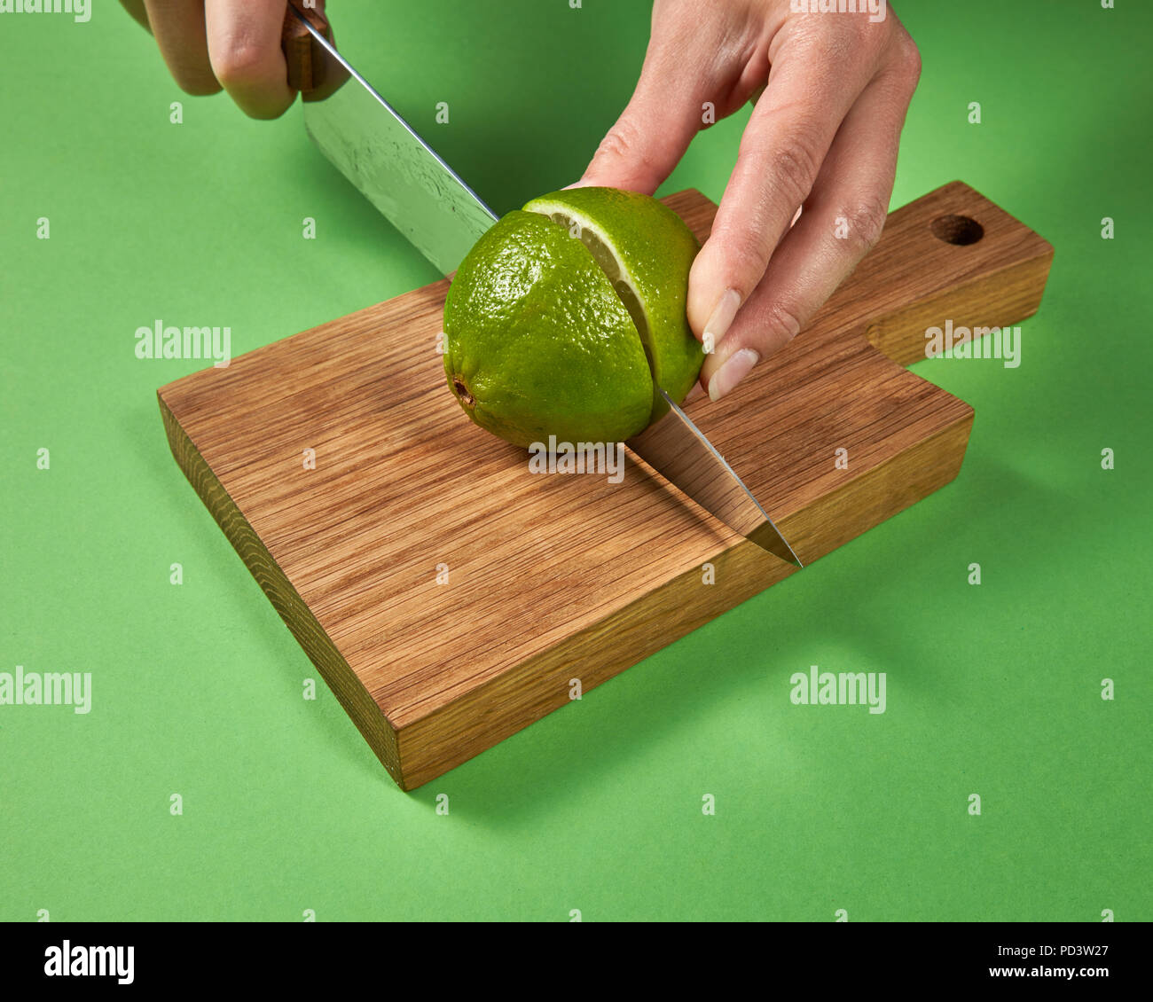 A girls' hands cut a green ripe lime in half on a wooden board on a green background. Stock Photo