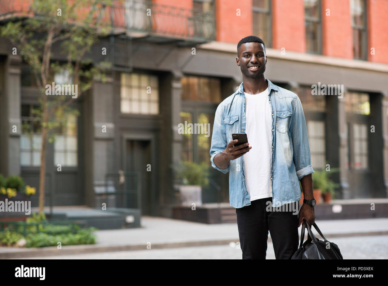 Young man with hand luggage holding cellphone in streets of New York, US Stock Photo
