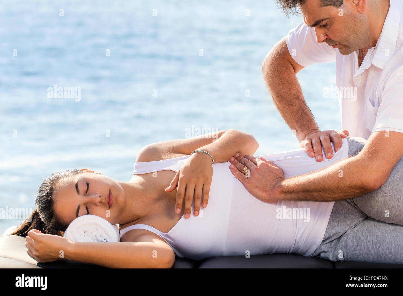 Close up portrait of male osteopath doing manipulative treatment on female hip. Therapist releasing lower back pain on woman against outdoor sea side  Stock Photo