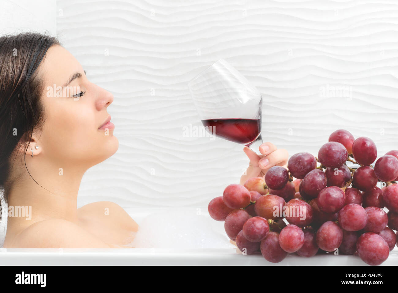 Close up portrait of young woman relaxing in foam bath with red wine and bunch of red grapes.Girl holding glass with red wine. Stock Photo