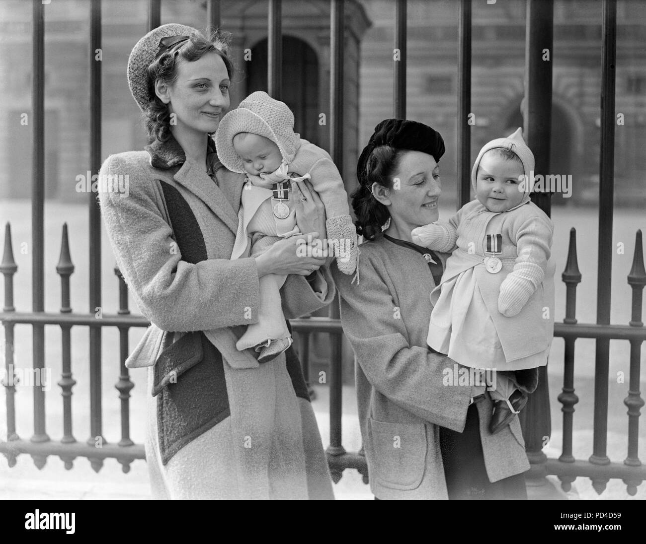 26th March, 1945. London, England. Posthumous Investiture at The palace. Mrs. May Kelly (left), of Kensington, London, whose husband won a second Bar to his D.S.M. lets her sleeping baby Mavis what the decoration. Also, Mrs. Margaret Barton, (right), of Liverpool, with her 8 month old daughter Brenda wearing her father's D.S.M. The awards were presented to the widows at an investiture at Buckingham Palace, when the King decorated next-of-kin of Empire heroes fallen in battle. Stock Photo