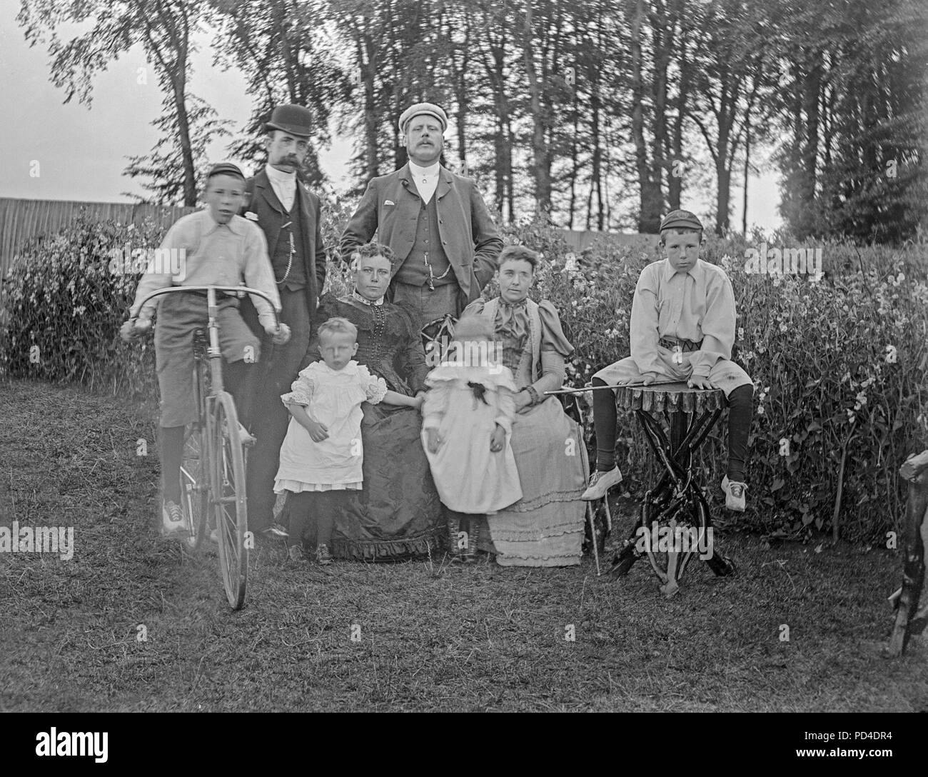 Alate Victorian photograph of a family posing for the camera, somewhere in England. There are two men and their wives, two young children, and two boys. One of the boys is sitting on a bicycle of the time. Stock Photo